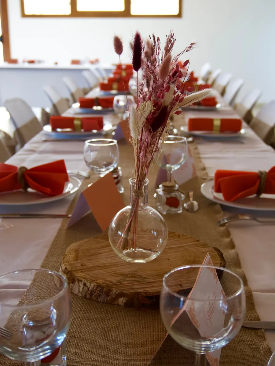 Longue table de fête dressée en perspective, chemin de toile de jute, serviettes corail, vase de fleurs séchées sur un rondin de bois et marque-place