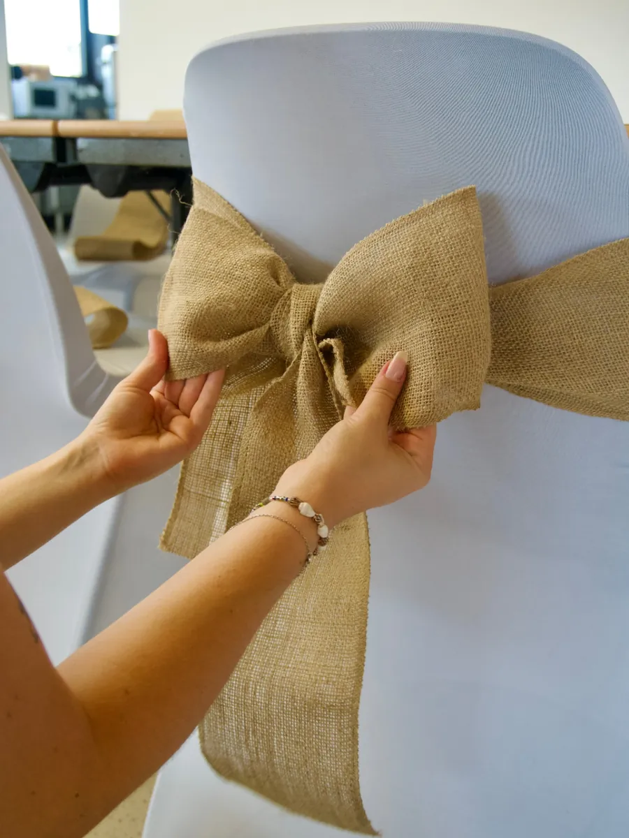 Mains nouant un large ruban de toile de jute sur une chaise habillée de blanc