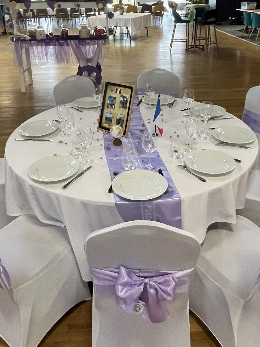 Table ronde dressée avec chemin de table lilas, chaises houssées de blanc à larges nœuds violets et buffeterie décorée de tulle