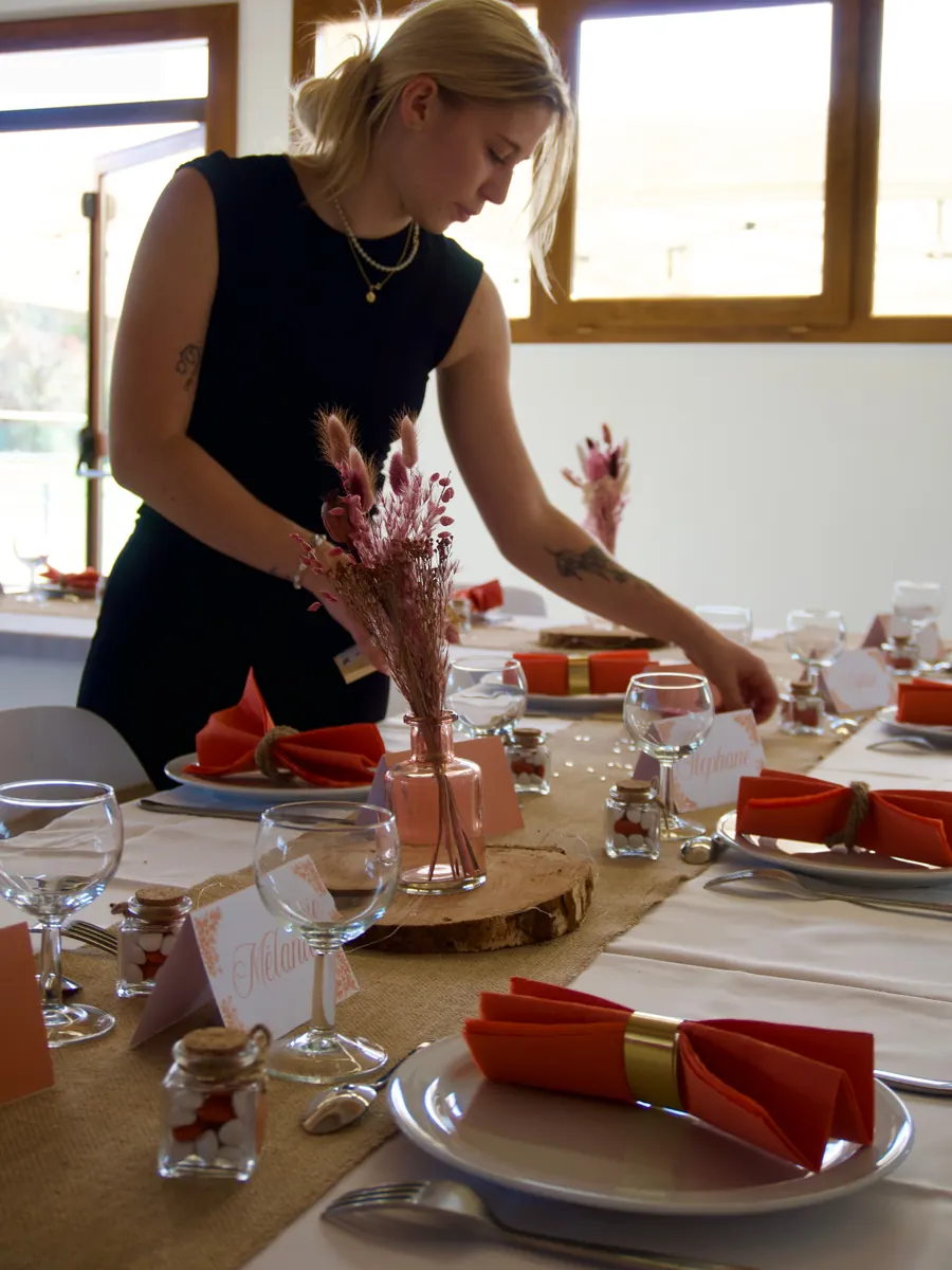 Justine en robe noire ajustant la décoration d'une longue table de fête, vases de fleurs séchées et marque-places alignés