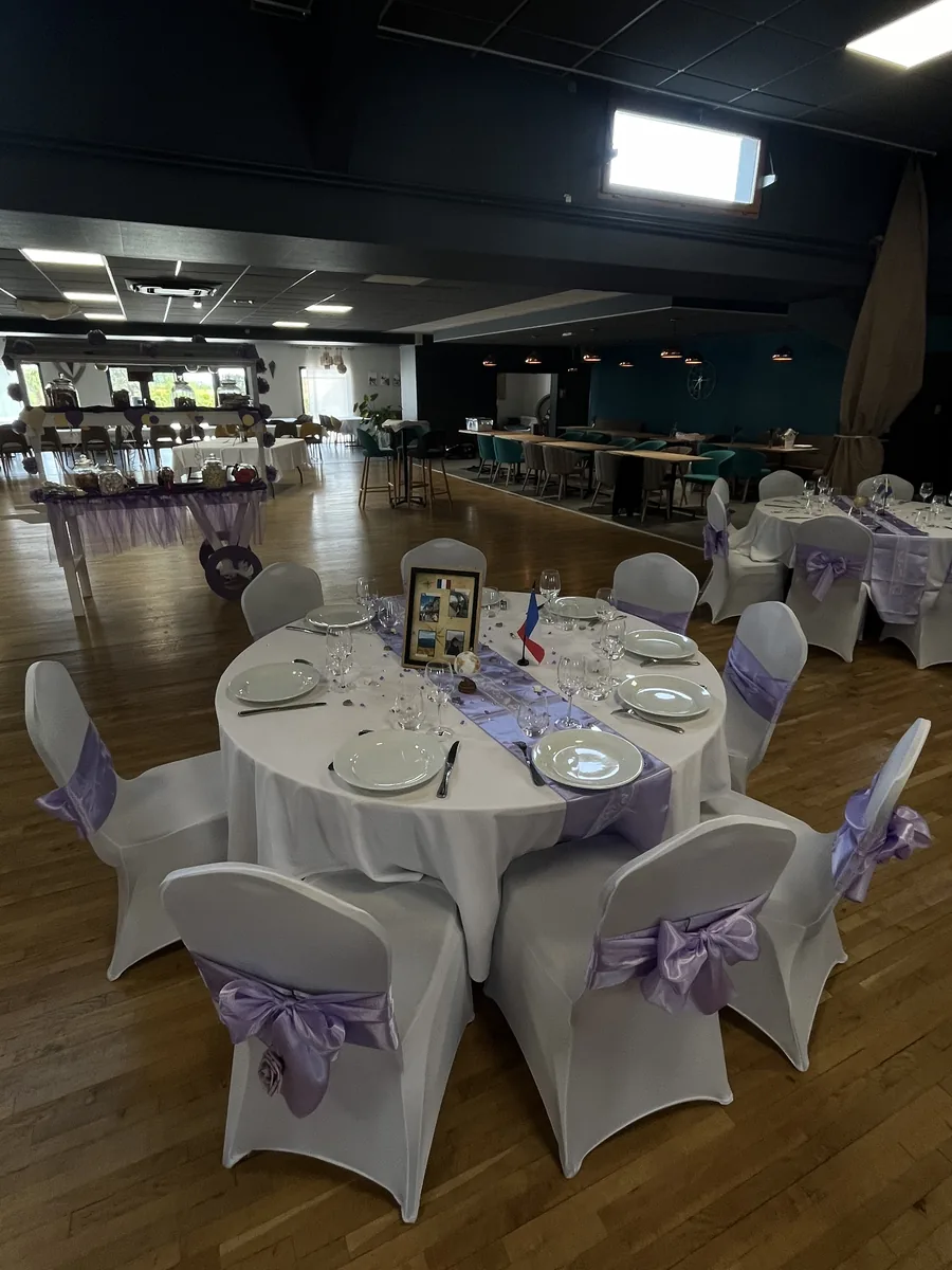 Salle de réception dressée pour un mariage : tables rondes en nappes blanches, chaises houssées de blanc avec nœuds lilas et chariot de douceurs