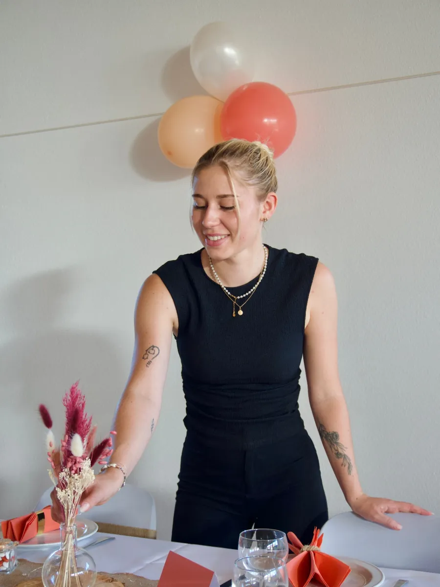 Justine disposant un petit vase de fleurs séchées sur une table dressée, ballons crème et corail au mur