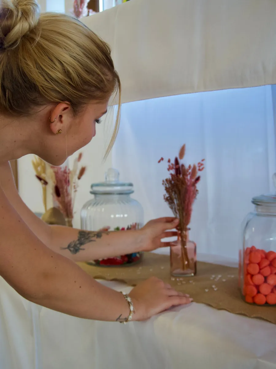 Justine dressant un buffet de bocaux en verre remplis de bonbons orange et un vase de fleurs séchées sur un chemin de toile de jute