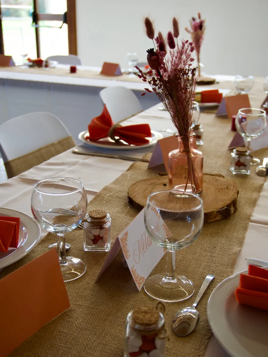 Détail d'une table dressée : fioles de dragées à bouchon de liège, rondin de bois portant un vase de fleurs séchées, serviettes terracotta pliées